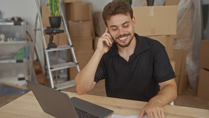 Man talking on phone while using laptop at table in building with packed boxes, ladder and drill visible; productivity moving day.