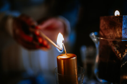 Individual lighting a golden candle with a matchstick, creating a warm glow in a dimly lit environment, enhancing the ambiance of a cozy evening setting