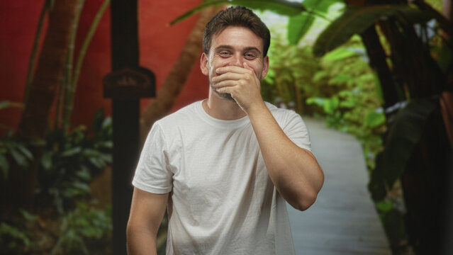 Man in white t shirt with hand covering mouth on a street walkway near plants, visible forearm and smile; amusement shyness. - Powered by Adobe