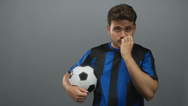 Young hispanic man in a blue and black soccer jersey holding a soccer ball and biting his nails with a direct gaze in a grey studio; anxiety. - Powered by Adobe