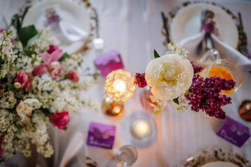 Elegant floral arrangement with peonies and lilacs on a beautifully set dining table, featuring candles and decorative elements for a festive celebration atmosphere