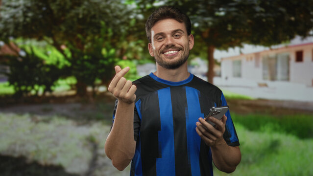 Young hispanic man smiling holds smartphone and makes finger heart gesture beside building in a green park; joy connection.