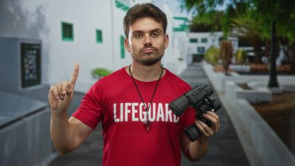 Man lifeguard wearing red tshirt with lifeguard print and whistle necklace points finger while...