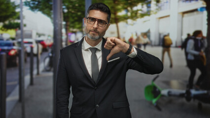 Fototapeta premium Man in business suit showing thumbs down with raised hand on street sidewalk near scooters and pedestrians; disapproval criticism.