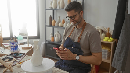 Man in denim apron holds smartphone and scrolls with his hands beside a pottery wheel and brushes...