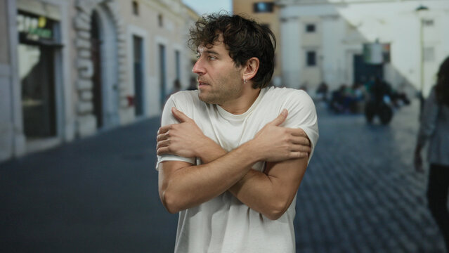 Hispanic man feeling cold outdoors in the street, wearing a white shirt, looking pensive as he embraces himself to keep warm in an urban setting with people and buildings.