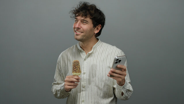Hispanic man in striped shirt holding ice cream and smartphone against grey background smiling with enjoyment highlighting modern casual lifestyle and technology interaction