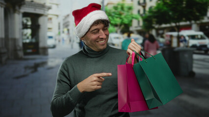 Hispanic man wearing christmas hat smiles while pointing to shopping bags on an outdoor street, capturing a festive urban shopping scene with vibrant colors and joyful expressions.