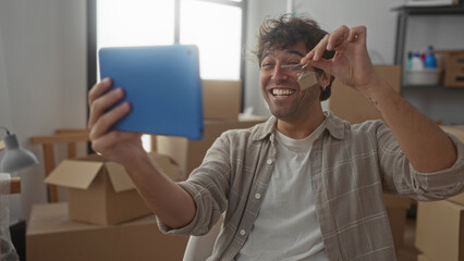 Hispanic man holding tablet and keys in new home surrounded by moving boxes joyfully video calling friends and family to celebrate moving day in bright living room