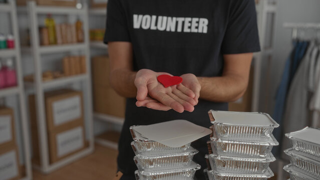 Hispanic man holding red heart symbol while smiling at camera in volunteer room surrounded by stacks of food trays exemplifying kindness and community support