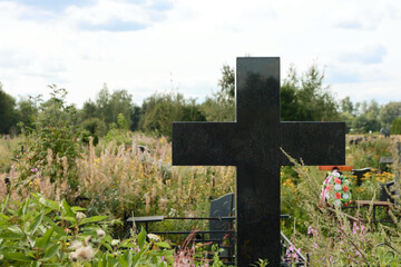 A Simple Polished Black Stone Cross Standing As A Tombstone In A Cemetery With Wildflowers And Tall Grass.