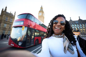 Fotobehang Londen rode bus Smiling african millennial woman taking a selfie in London with a red double-decker bus and Big Ben in the background. Travel portrait that conveys joy, sightseeing and city energy.  © CarlosBarquero