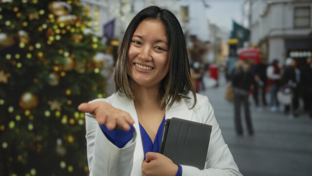 Woman smiling outdoors in a city street holding a tablet with festive decorations and reaching out with a friendly gesture.