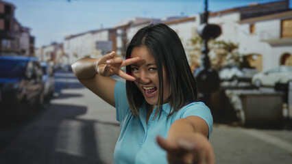 Fototapeta premium Chinese woman making a playful victory gesture on a city street pointing towards the camera with a lively expression and urban background.