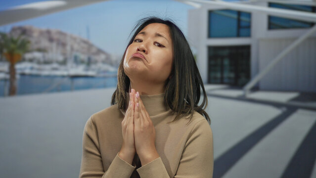 Woman in beige sweater with hands clasped in front of modern seaside building backdrop showing expression of pleading by the sea at a port with boats visibly docked near.