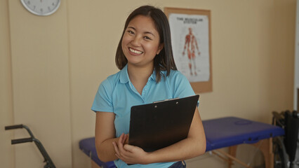 Woman smiling holding clipboard in hospital clinic room with anatomical poster clock visible on wall portraying cheerful healthcare professional in indoor workplace setting