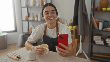 Woman potter in apron smiles while holding ceramic mug and checks red smartphone in studio workspace; creative joy.