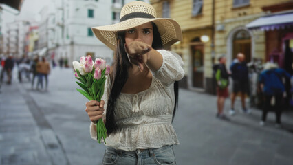 Woman holding pink tulips and pointing finger at camera on a crowded european cobbled street with shopfronts; confidence.