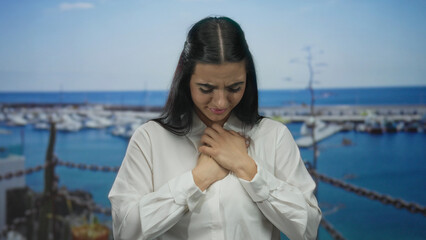 Fototapeta premium Woman with hands on chest standing at a port with boats in the background, wearing a white shirt, demonstrating an emotional and reflective moment outdoors by the sea.