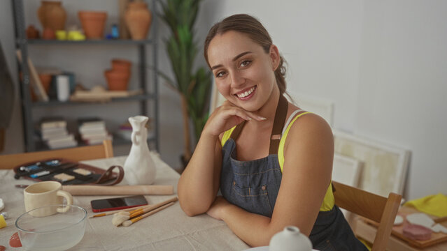 Woman with bare arm and denim apron resting hand on chin at artisan studio table with brushes and pottery; creative calm.
