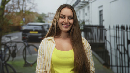 Woman smiles at camera wearing yellow tanktop and open striped shirt on a city street with bicycles and parked car; confidence warmth.