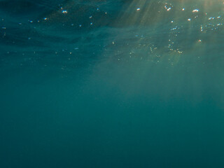 Fototapeta premium Dark blue ocean surface seen from underwater. Abstract waves underwater and rays of sunlight shining through, Sun light rays undersea deep, Underwater background with sea bottom, Mediterranean sea.