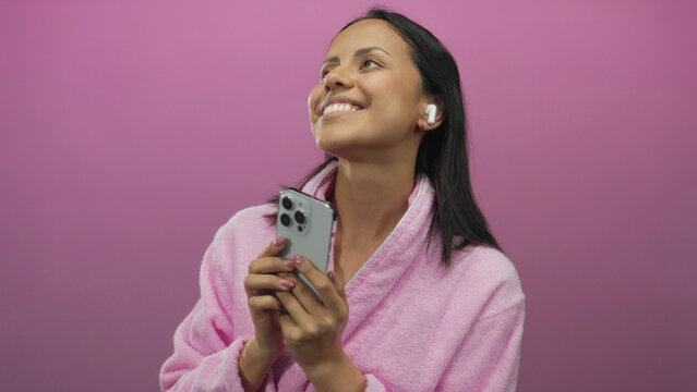 Young woman in a pink bathrobe enjoys music with earbuds and smartphone against an isolated vibrant pink background, exuding joy and relaxation.