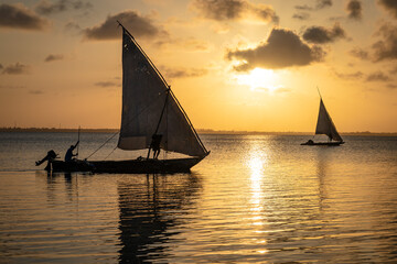 Two traditional sailboats are silhouetted against a serene, golden sunset on calm, reflective water