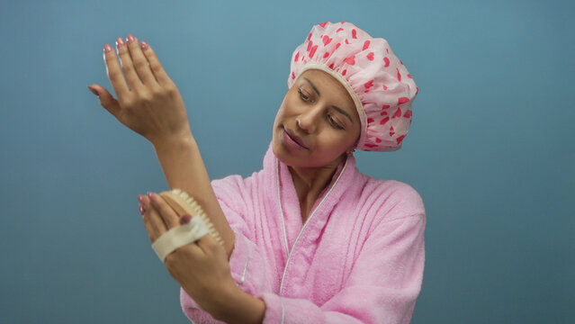 Hispanic woman in pink bathrobe and heart shower cap exfoliating arm with brush against blue isolated wall, showcasing skincare self-care routine beauty. - Powered by Adobe