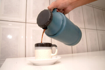 Hand pouring hot coffee from a blue thermos into a speckled ceramic mug on a white kitchen counter. Perfect for breakfast, lifestyle, or beverage content.