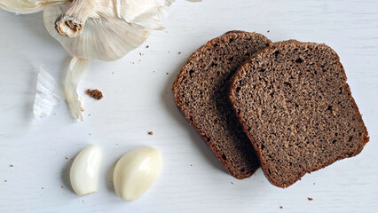 Two slices of dark rye bread, head of garlic, and two garlic cloves on white wooden surface. Ingredients for healthy snack or meal preparation for recipe blogs, cookbooks, health-conscious content.