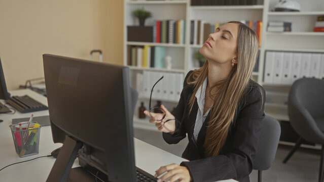 Young hispanic woman adjusting glasses and typing at desktop computer while sitting at an office desk with shelves behind; concentration productivity determination.