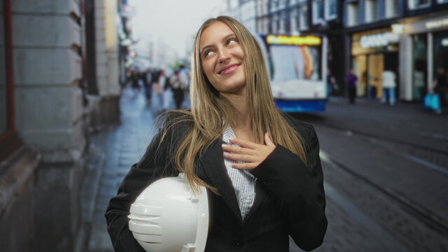 Young woman holding white hardhat with hand on chest smiling on busy street with tram and shops in view; career pride.