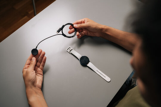 Top view from shoulder of unrecognizable man holding smartwatch inductive charger, preparing to power up smart watch, sitting at table. Concept of healthy lifestyle.