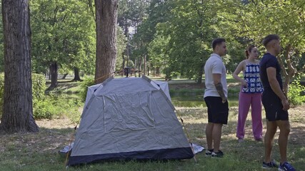 A group of young friends prepares for a fun outdoor adventure, setting up a tent in a beautiful, serene park. They enjoy nature and spending quality time together, embracing the camping experience.