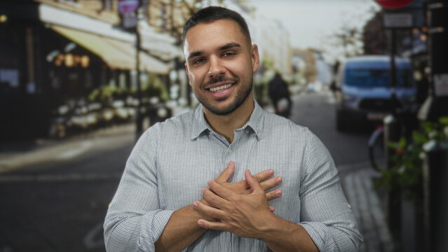 Smiling hispanic man in striped shirt crosses hands over chest on sunlit street; gratitude sincerity.
