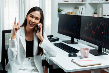 Young beautiful Asian businesswoman talking to broker on phone with looking on pc screen to analyze...