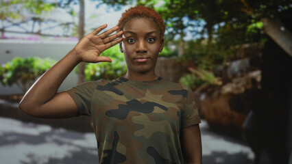 Woman in camouflage uniform salutes with hand to head under trees in green sunlit park outdoors; duty.