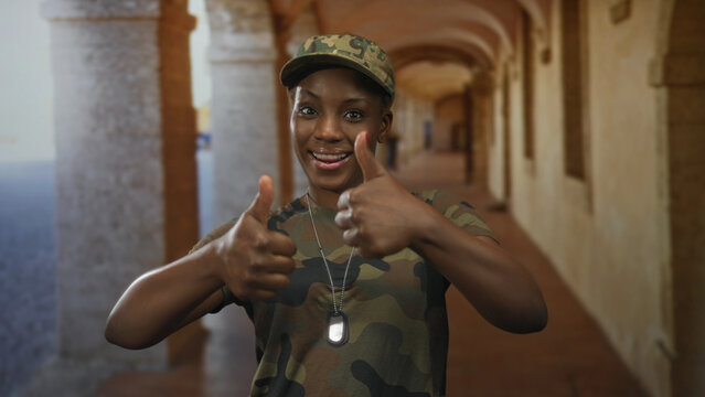 Young african american woman soldier in camouflage uniform gives thumbs up gesture in stone building corridor; approval. - Powered by Adobe