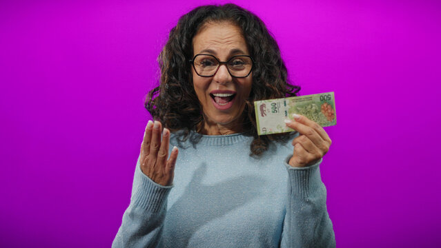 Middle-aged woman holding argentine pesos with a puzzled expression stands against a vibrant purple wall, suggesting themes of finance and confusion.