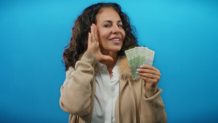 Middle-aged woman smiling and holding romanian banknotes against a solid blue background, conveying happiness and financial success with an isolated joyful expression.