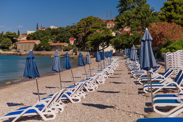 Row of empty loungers and blue umbrellas on a quiet pebble beach with calm water. Concept of summer vacation design, travel marketing and coastal background.