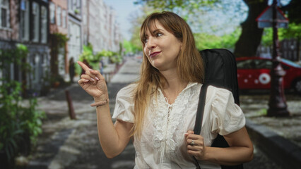 Fototapeta premium Woman points finger upward on a sunlit city street holding backpack strap and smiling while looking to the side; travel confidence playful.