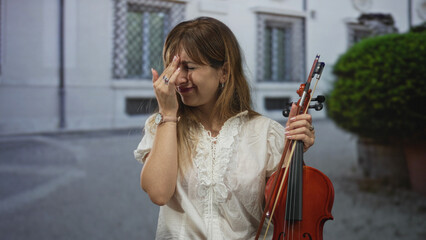 Woman violinist holding violin and bow with hand to forehead in front of building on street  frustration exhaustion. © Krakenimages.com
