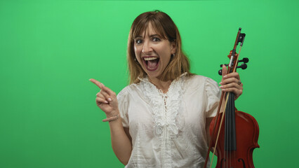Woman holding violin and points finger to right in green screen studio, smiling with open mouth; excited performance.