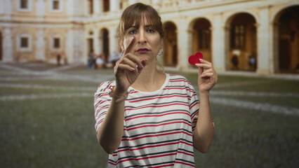 Woman points finger forward holding small red felt heart beside arches of old town building...