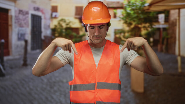 Man engineer in orange safety vest and hardhat pointing both index fingers down on street; safety professionalism.