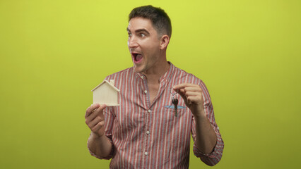 Man holding a wooden house model and metal keys, mouth open in surprise, wearing striped shirt in a lime green studio; home buying excitement.