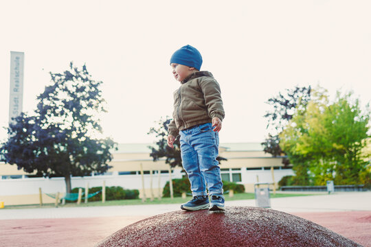 Child joyfully climbing a rubber mound at a playground in autumn, surrounded by trees and a school building, capturing the essence of childhood playtime and adventures - Powered by Adobe