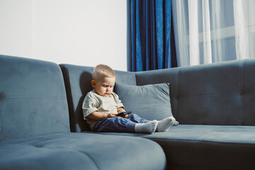 Curious toddler explores smartphone while sitting comfortably on a blue couch in a sunlit living room